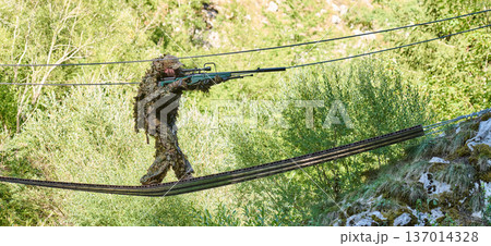 Camouflaged Sniper Soldier Crossing Narrow Rope Bridge In Forested Mountain Terrain On Patrol Camouflaged Sniper Soldier Crossing Narrow Rope Bridge In Forested Mountain Terrain On Patrol 137014328