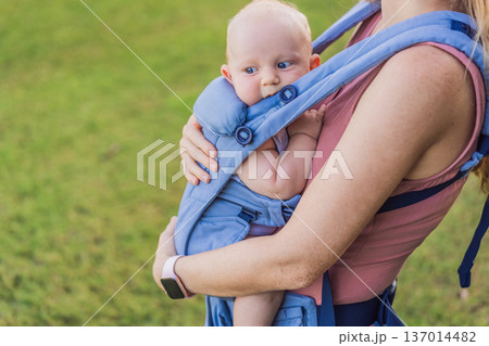 Mother using ergonomic baby carrier during a walk with her infant outdoors. Comfortable babywearing, active motherhood lifestyle, bonding, modern parenting, hands free care and everyday family routine 137014482