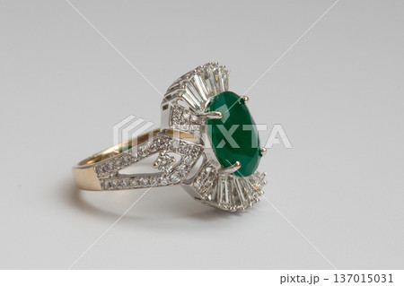 A gold ring featuring an oval cut green emerald surrounded by baguette cut diamonds on a clean white background. A gold ring featuring an oval cut green emerald surrounded by baguette cut diamonds on a clean white background. 137015031