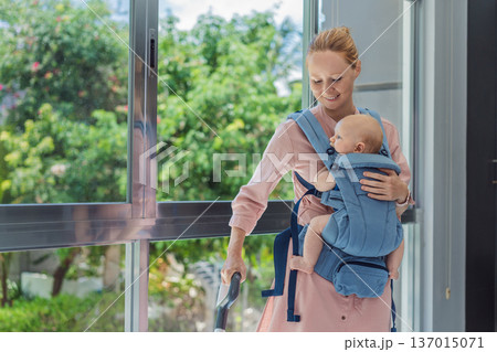 Woman vacuuming a room while carrying her baby in an ergonomic carrier. Modern motherhood, multitasking parent, home chores with infant, balance of childcare and housework concept. 137015071