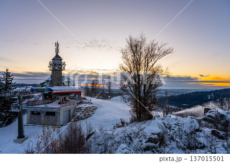 Snow covers the ground around the Tanvaldsky Spicak lookout tower. The winter landscape reveals distant mountains and a soft sunset. Visitors may enjoy the views from this resort area. Snow covers the ground around the Tanvaldsky Spicak lookout tower. The winter landscape reveals distant mountains and a soft sunset. Visitors may enjoy the views from this resort area. 137015501