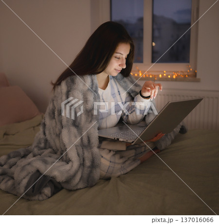 Girl sits on bed wrapped in blanket using laptop as night, writing messages and browsing on in internet 137016066