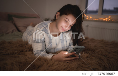 Young girl lies on a bed and looks at her phone during the evening, surfing on internet before sleep 137016067