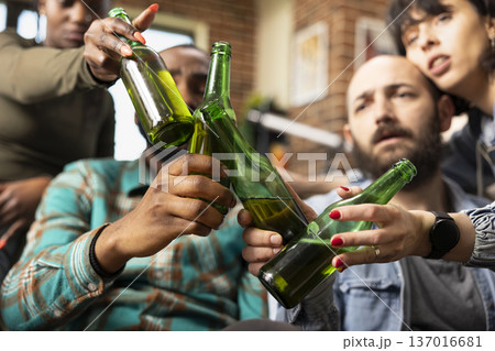 Close up of diverse friends raising beer bottles in toast, celebrating together in cozy living room with brick wall. Happy multiethnic group bonding, sharing drinks and enjoying casual gathering. Close up of diverse friends raising beer bottles in toast, celebrating together in cozy living room with brick wall. Happy multiethnic group bonding, sharing drinks and enjoying casual gathering. 137016681