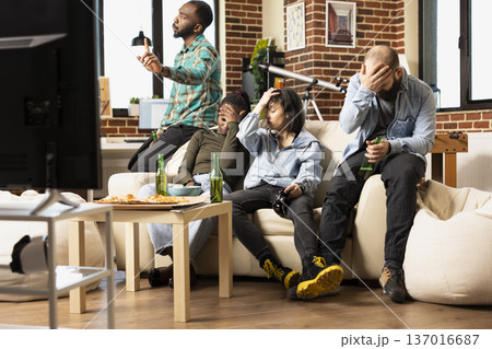 Frustrated friends sit with head in hand after losing game, showing disappointment and annoyance. Group of young adults in casual apartment reacts with sadness after tournament defeat. 137016687