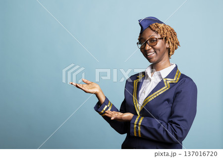 Cheerful air hostess stands against blue background, gesturing toward blank mockup space. Female flight attendant promotes airline offers, travel deals and professional aviation services in studio. 137016720