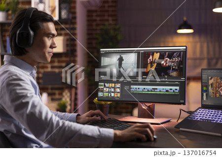 Asian filmmaker with headphones editing video footage on computer in home office. Creative male freelancer works on cinematic project, applying color grading and visual effects for film production. 137016754