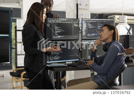 Male and female software developers stand by computer monitors while brainstorming coding project for startup. Diverse team collaborate on refining HTML scripts and building modern website together. 137016792