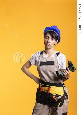 Disappointed repairwoman holds power drill, noticing mistakes on renovation site. Unhappy female builder places one hand on waist and grips tool, appearing worried after mishap during construction. 137016794