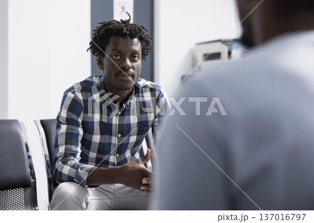 Young black patient sits calmly in modern hospital lobby while waiting for medical checkup. African american man looks directly at camera, presenting relaxed, confident moment before his consultation. Young black patient sits calmly in modern hospital lobby while waiting for medical checkup. African american man looks directly at camera, presenting relaxed, confident moment before his consultation. 137016797