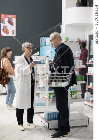 Woman pharmacist stands beside elderly male customer, explaining healthcare options near store shelves. The interaction highlights trust, expertise and personalized service in clean, modern pharmacy. Woman pharmacist stands beside elderly male customer, explaining healthcare options near store shelves. The interaction highlights trust, expertise and personalized service in clean, modern pharmacy. 137016814