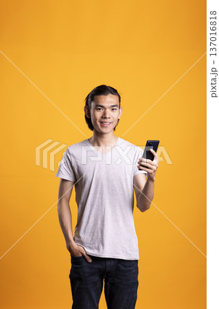 Happy male model in casual outfit poses with one hand in pocket, holding smartphone and looking at camera in modern studio. Young man texting friends on mobile device and browsing social media. 137016818