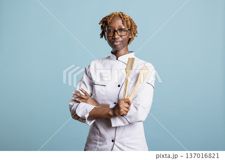 Focused female chef stands with arms crossed while holding wooden cooking utensils in studio. Young culinary professional expressing confidence, skill and discipline in food industry. 137016821