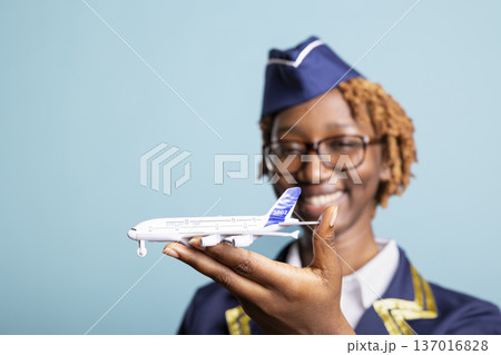 Joyful female airline employee in uniform holding miniature airplane and smiling at camera. Professional cabin crew member promotes aviation industry and passion for global travel. Joyful female airline employee in uniform holding miniature airplane and smiling at camera. Professional cabin crew member promotes aviation industry and passion for global travel. 137016828