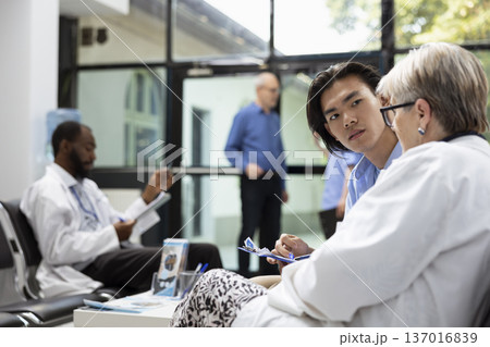 Asian man meets with caucasian physician in clinic lobby before medical testing. Senior doctor reviews procedures and documents, highlighting preparation, trust and professional healthcare guidance. 137016839
