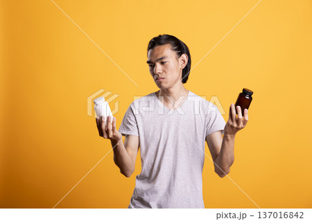 Man examines two different pill bottles, feeling unsure which medication to choose. Person compares white and brown containers with confused expression, showing decision making in personal health care Man examines two different pill bottles, feeling unsure which medication to choose. Person compares white and brown containers with confused expression, showing decision making in personal health care 137016842