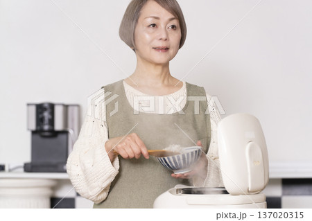 キッチンで炊飯器から炊きたてご飯を茶碗によそうシニア女性 137020315