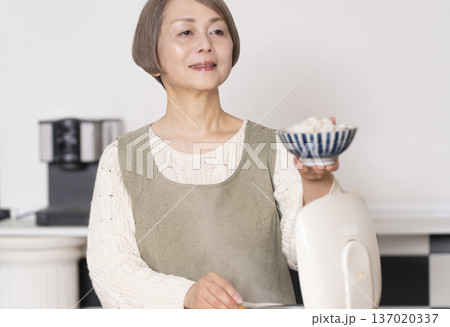 キッチンで炊飯器から炊きたてご飯を茶碗によそうシニア女性 137020337
