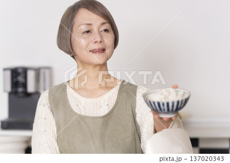 キッチンで炊飯器から炊きたてご飯を茶碗によそうシニア女性 137020343