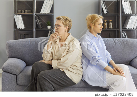 Mature mother and adult daughter sitting back-to-back with serious expressions Mature mother and adult daughter sitting back-to-back with serious expressions 137022409
