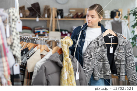 Interested woman standing in clothing shop and looking new coat for autumn-winter season. Shopping and seasonal sales concept Interested woman standing in clothing shop and looking new coat for autumn-winter season. Shopping and seasonal sales concept 137023521