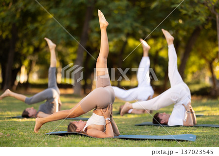 Athletic young girl demonstrating flexibility in modified Shoulder Stand Eka Pada Sarvangasana during group yoga training on green meadow in summer park Athletic young girl demonstrating flexibility in modified Shoulder Stand Eka Pada Sarvangasana during group yoga training on green meadow in summer park 137023545