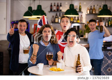 Happy young adult fans waving British flag while drinking beer and watching match together in sports bar Happy young adult fans waving British flag while drinking beer and watching match together in sports bar 137023689