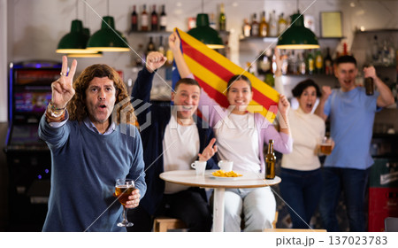 Company of emotional young adult fans supporting favorite team of Estelada with flag while resting in sports bar Company of emotional young adult fans supporting favorite team of Estelada with flag while resting in sports bar 137023783