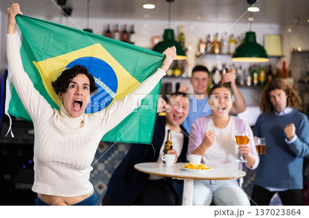 Company of enthusiastic young adult sports fans waving flag of Brazil and supporting national team with beer in pub 137023864