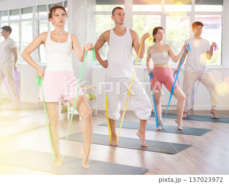 Positive woman with sporty women and men engaged in group class standing and perform exercise with resistance bands in pilates studio Positive woman with sporty women and men engaged in group class standing and perform exercise with resistance bands in pilates studio 137023972