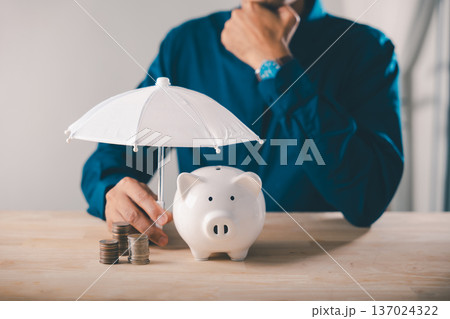 Bank piggy money protect financial insurance concept risk umbrella collage finance shown as man covers coins with white umbrella for long term investment safety on wooden work desk Bank piggy money protect financial insurance concept risk umbrella collage finance shown as man covers coins with white umbrella for long term investment safety on wooden work desk 137024322