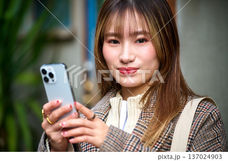 スマートフォンを操作する20代日本人女性 チェック柄の冬服でお出かけ ライフスタイル ポートレート スマートフォンを操作する20代日本人女性 チェック柄の冬服でお出かけ ライフスタイル ポートレート 137024903