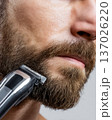 Extreme close-up of adult middle aged man trimming brown beard with professional electric clipper. Detailed male grooming concept. Facial hair maintenance, barbering, and hygiene. Bright studio light. 137026220