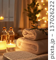 Two soft white rolled towels lie next to glass essential oil bottles and burning candles in a dimly lit room, evoking a sense of cozy luxury self-care and evening relaxation at a home SPA. 137026222