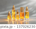 Five glass cosmetic dropper bottles with golden caps and amber liquid stand in a row on a reflective surface, illuminated by soft direct light and shadows for a minimalist luxury serum or oil mockup. 137026230