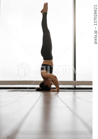 A woman in black athletic wear performs a headstand yoga pose on a mat in a bright room with large windows and wooden floors A woman in black athletic wear performs a headstand yoga pose on a mat in a bright room with large windows and wooden floors 137026621