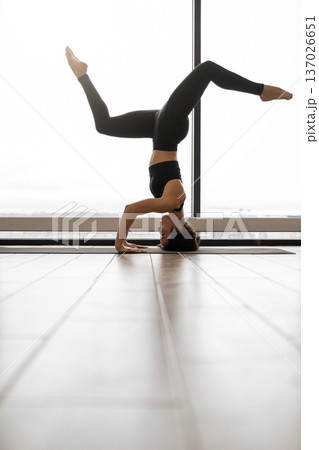 A woman performs a headstand yoga pose with legs extended in a bright, modern studio with large windows and wooden floors 137026651