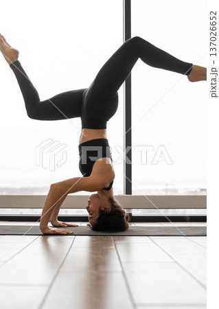A woman in black activewear performs a challenging yoga headstand pose with her legs extended upwards against a bright window backdrop A woman in black activewear performs a challenging yoga headstand pose with her legs extended upwards against a bright window backdrop 137026652