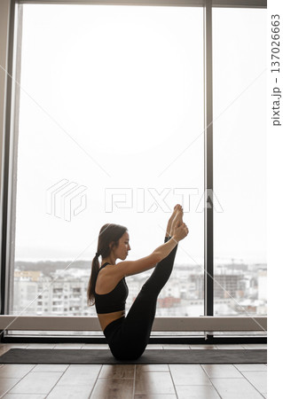 A woman in black activewear performs a yoga pose, balancing on her mat with legs extended towards the ceiling, framed by a large window overlooking a city skyline A woman in black activewear performs a yoga pose, balancing on her mat with legs extended towards the ceiling, framed by a large window overlooking a city skyline 137026663