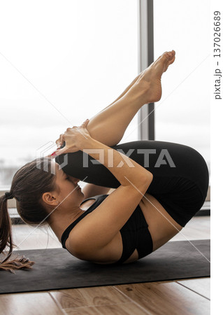 A woman in black activewear performs a yoga pose on a mat, drawing her knees towards her chest A woman in black activewear performs a yoga pose on a mat, drawing her knees towards her chest 137026689