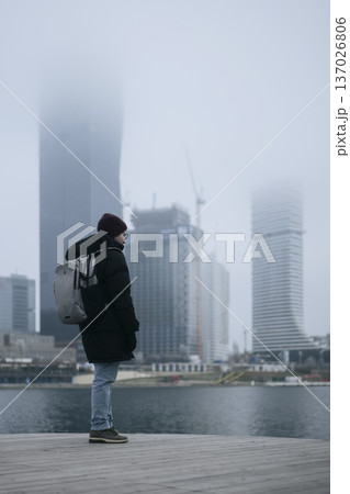 Man in winter coat overlooking a foggy city waterfront. Concept of a city winter activity Man in winter coat overlooking a foggy city waterfront. Concept of a city winter activity 137026806