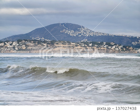 Calm waves meet mountain horizon, Tranquil seaside views featuring rugged mountains and overcast sky 137027438