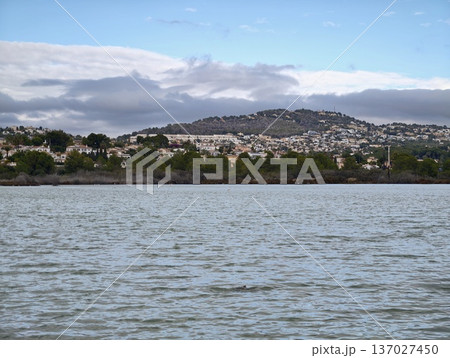 Quiet coast with distant mountain settlement, Tranquil waters surrounded by muted sky and rolling hills Quiet coast with distant mountain settlement, Tranquil waters surrounded by muted sky and rolling hills 137027450