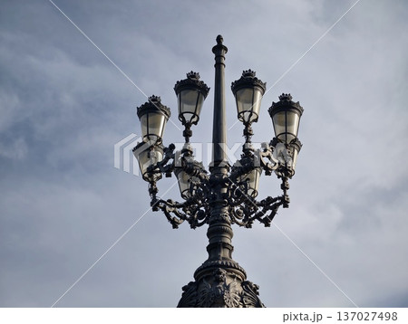 Decorative streetlight under cloudy sky, Ornate cast iron lamppost with multiple lanterns overhead Decorative streetlight under cloudy sky, Ornate cast iron lamppost with multiple lanterns overhead 137027498