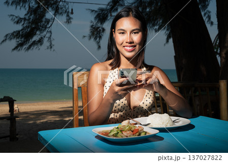 Smiling Woman Using Smartphone While Dining at Beachside Cafe 137027822