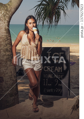 Smiling woman holding ice cream cone while standing on sandy beach near sign full length shot Smiling woman holding ice cream cone while standing on sandy beach near sign full length shot 137027823