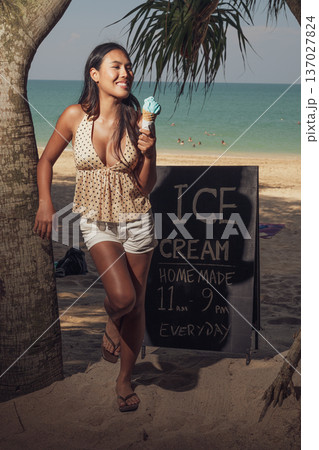 Smiling woman holding ice cream cone while standing on sandy beach near sign full length shot 137027824