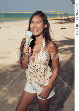 Smiling woman holding ice cream cone on sandy beach with volleyball court behind Smiling woman holding ice cream cone on sandy beach with volleyball court behind 137027844