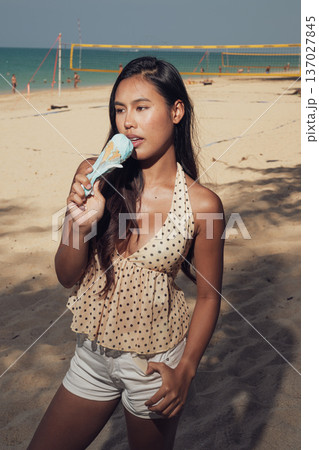 Smiling woman holding ice cream cone on sandy beach with volleyball court behind Smiling woman holding ice cream cone on sandy beach with volleyball court behind 137027845
