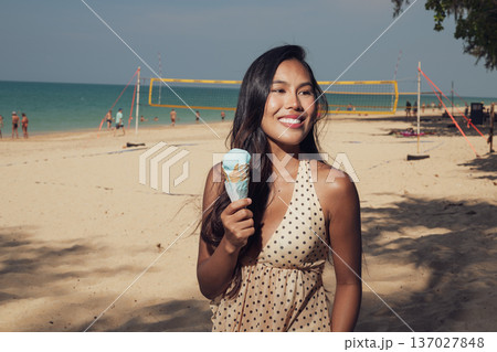 Smiling woman holding ice cream cone on sandy beach with volleyball court behind Smiling woman holding ice cream cone on sandy beach with volleyball court behind 137027848